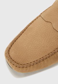 Zapato de cuero color marrón claro con una superficie texturizada, que presenta costuras en contraste a lo largo del borde. Punta redondeada y un diseño suave y flexible.