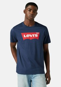 Joven vistiendo una camiseta de Levi's de color azul marino con un logo rojo y blanco, combinada con jeans de color azul claro, de pie frente a un fondo liso.