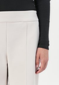 Beige broek gemaakt van zachte stof, met een brede tailleband, twee voorplooien en een schone, op maat gemaakte silhoeët. Donkere top zichtbaar aan de zijkant.