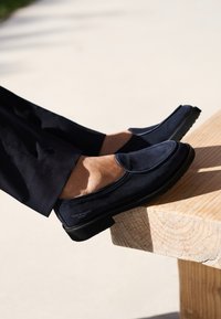Navy blue suede loafers with a rubber sole, featuring a rounded toe and stitched detailing along the upper. Worn with black pants.