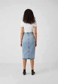 Fustă din denim, tip creion, de culoare bleu deschis, cu margine brută, închidere cu fermoar și o etichetă de marcă pe spate. Poartă un tricou alb simplu.