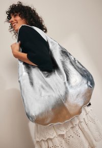 Nagy ezüst fémes bőr táska sima textúrával, laza formával. Széles nyílással és puha szélekkel rendelkezik.