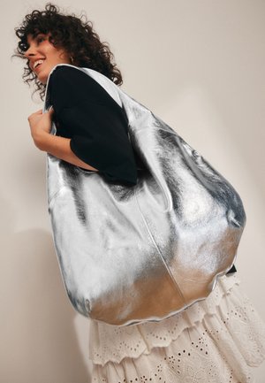 Didelis sidabrinis metalinis odinis krepšys su lygiu paviršiumi, turintis laisvos formos siluetą. Yra platus atidarymas ir minkšti kraštai.