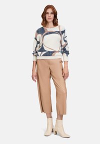 Pullover in Creme mit abstrakten Mustern in Marineblau und Beige; weicher, strukturierter Stoff. Hellbraune, weit geschnittene Lederhosen; cremefarbene Stiefeletten mit Blockabsatz.