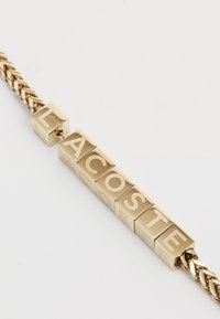 Bracelet en chaîne dorée avec des blocs cubiques portant les lettres "LACOSTE" embossées à la surface. La chaîne présente un design tressé.