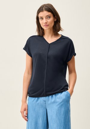 Dark navy short-sleeve blouse with a V-neck, subtle stitching detail, and relaxed fit. Worn with light blue, loose-fitting trousers.