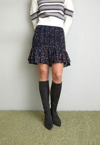 Femme portant une jupe à volants fleurie bleu marine, un pull blanc à motifs et des bottes hautes en suède noires, debout sur un tapis vert.