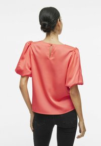 Blusa in satin corallo con maniche a sbuffo e apertura a occhio di bottone sulla schiena; texture liscia e vestibilità rilassata, abbinata a jeans neri.