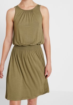 Nainen pukeutuneena hihattomaan oliivinvihreään mekkoon, jossa on koota kaulus ja joustava vyötärö, seisoo yksiväristä valkoista taustaa vasten.