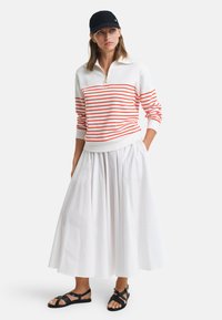 Pull zip blanc avec des rayures rouges, associé à une jupe fluide blanche et des sandales noires. L'ensemble est complété par une casquette décontractée.