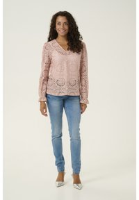 Donna con capelli ricci che indossa una blusa rosa in pizzo a maniche lunghe, jeans blu e décolleté argentati a punta, in piedi su uno sfondo chiaro.