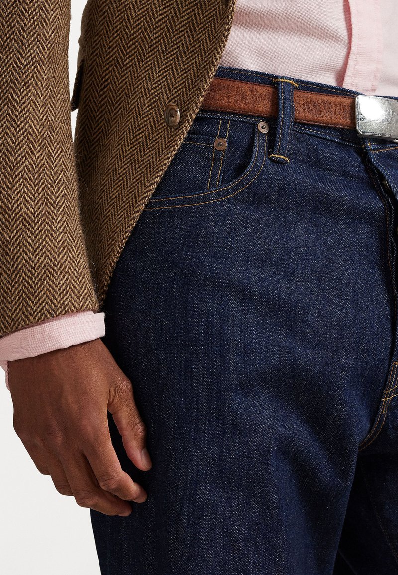 Brown herringbone blazer, light pink shirt cuff, dark blue denim jeans, brown leather belt with silver buckle, casual outfit detail.