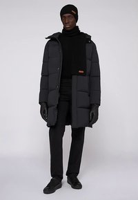 Abrigo acolchado en gris oscuro de diseño hasta la rodilla, combinado con una bufanda, guantes y gorro negros. Pantalones y zapatos negros completan el conjunto.