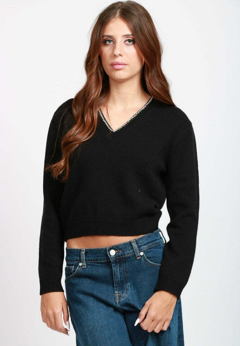 Giovane donna con lunghi capelli castani che indossa un maglione nero a scollo a V corto e jeans blu a vita alta su uno sfondo semplice.