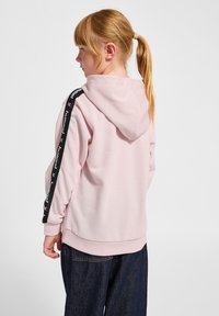 Roze hoodie met een zwarte logo-stripe langs de mouwen, voorzien van een capuchon en geribbelde manchetten; gedragen met donkere spijkerbroek.