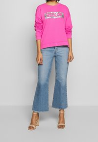 Sudadera fucsia con gráfico metálico plateado "REPLY", corte relajado y puños acanalados. Combinada con jeans acampanados de color azul claro y sandalias de tacón nude.