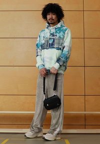 Felpa con cappuccio con motivi astratti blu e verdi, abbinata a pantaloni di velluto grigi con strisce bianche e una piccola borsa a tracolla nera.