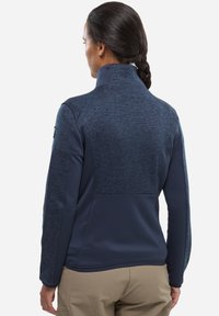 Veste en polaire bleu marine avec col montant, comportant un panneau arrière texturé et des manches lisses. Des accents cousus mettent en valeur le design.