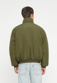 Olive green puffer jacket with a high collar, elastic waistband, and soft texture. Features a relaxed fit and smooth fabric.