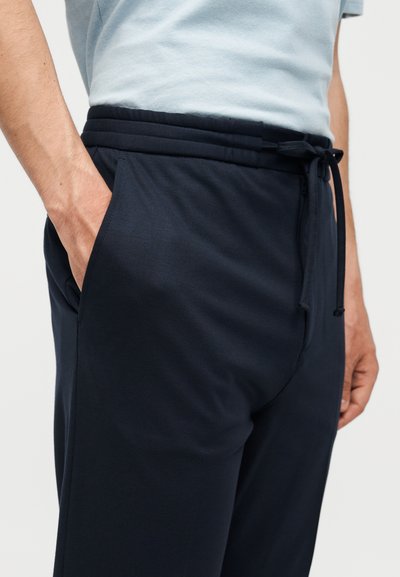 Κοντινό πλάνο ενός ατόμου που φοράει μπλε παντελόνια με κορδόνι, με τα χέρια στις τσέπες και ένα ανοιχτό μπλε πουκάμισο κεκλιμένο μέσα.