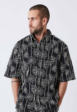 Homme aux cheveux courts et barbe portant une chemise noire à manches courtes avec un motif floral brodé blanc, regardant vers la gauche avec les mains dans les poches.
