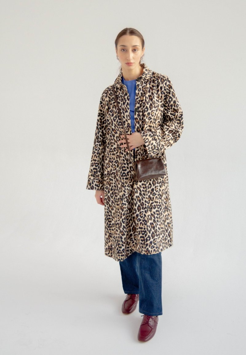 Cappotto lungo con stampa leopardata e colletto, realizzato in tessuto leggero, con tasche laterali; indossato sopra una maglietta blu con jeans scuri e scarpe borgogna.