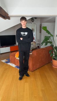 Mladý muž v čiernom oblečení stojí so založenými rukami v modernom obývacom priestore s oranžovou pohovkou, televízorom a veľkou izbovou rastlinou na drevenej podlahe.