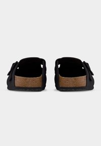 Ζευγάρι μαύρων σανδαλιών τύπου slip-on με πάτους από φελλό, ρυθμιζόμενα λουριά και υφασμένα ελαστικά πέλματα, εμφανιζόμενα από πίσω σε λευκό φόντο.