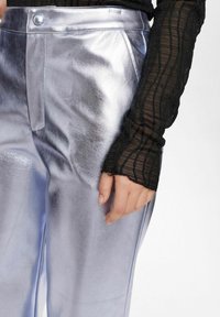 Pantaloni metallici d'argento con una texture liscia, dotati di chiusura con bottone e cuciture laterali. Una maglietta nera trasparente a maniche lunghe con texture è parzialmente visibile.