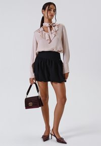 Donna che indossa una blusa a volant di un leggero rosa, una gonna nera alta in vita, tacchi bordeaux e tiene una borsa bordeaux con accessori dorati.