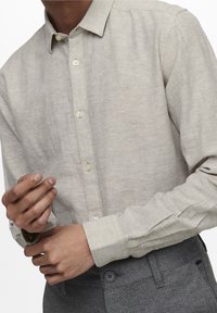 Uomo che sistema il polsino di una camicia button-up beige, indossando pantaloni grigi con bottone visibile e passanti per la cintura.