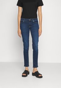 Dark blue jeans with a slim fit, featuring a mid-rise waist, five-pocket design, and contrasting stitching. Worn with a black t-shirt and black sandals.