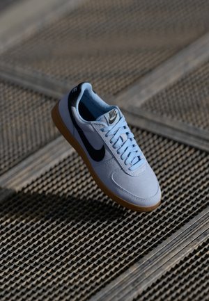 Baskets blanches Nike avec un swoosh noir et une semelle en gomme, flottant au-dessus d'une surface en grille métallique texturée sous éclairage extérieur.