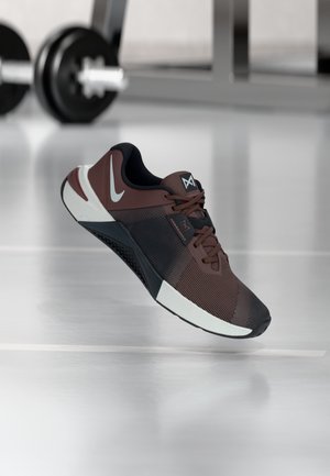 Zapatilla deportiva marrón y negra con suela blanca y cordones flotando sobre el suelo del gimnasio, con mancuernas desenfocadas al fondo.