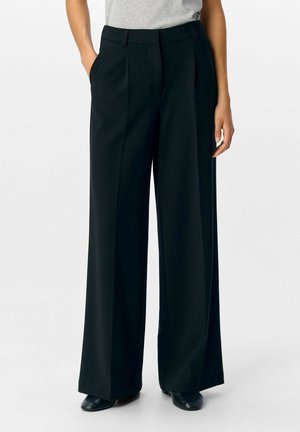 Schwarze, weit geschnittene Hosen aus glattem Stoff, mit hoher Taille, vorderen Falten und seitlichen Taschen, kombiniert mit einem enganliegenden grauen Oberteil.