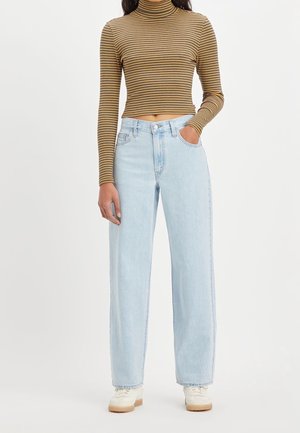 Kvinde iført lyseblå vidde jeans, beige og sort stribet langærmet rullekrave crop top og hvide sneakers, stående med den ene hånd i lommen.