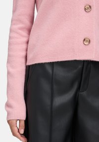 Nahaufnahme einer Person, die einen rosa Wollpullover mit Holzknöpfen und schwarze Lederhosen trägt, die Hand entspannt an der Seite.