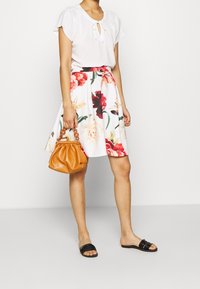 Vit blus med nyckelhålsdetalj, blommig knälång kjol i röda, rosa och gröna nyanser, svarta slip-on sandaler och en tan-handväska.