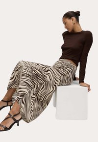 Hnědý top s dlouhým rukávem v kombinaci se smetanovou sukní s hnědým zebřím vzorem. Modelka sedí na bílém bloku a má na sobě černé sandály na podpatku.