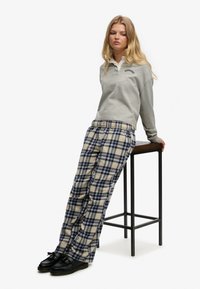 Jeune femme appuyée sur un tabouret haut portant un sweat-shirt gris, un pantalon à carreaux bleu et beige, et des chaussures noires, sur fond blanc.