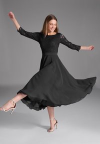 Robe noire avec un corsage en dentelle et une jupe fluide. La jupe présente un design superposé, associée à des sandales à talons hauts argentées.
