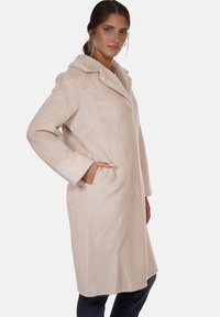 Cappotto lungo beige con cappuccio, caratterizzato da una texture morbida, tasche laterali e un colletto sagomato. Indossato sopra jeans scuri.