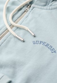 Lichtblauwe hoodie met een zachte textuur, voorzien van een zilverkleurige ritssluiting, off-white trekkoorden en "SUPERDRY" geborduurd in blauw.