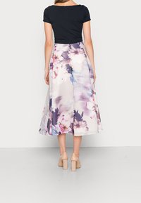 Robe midi florale avec un haut noir ajusté et une jupe plissée de couleur claire ornée de fleurs violettes et roses. Des talons transparents complètent le look.