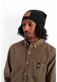 Black knit beanie with a brown leather patch. Olive green corduroy shirt with yellow buttons and a black logo patch on the pocket.