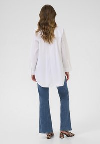 Camicia bianca in cotone a maniche lunghe con orlo curvo, abbinata a jeans svasati blu e mule con tacco in stampa leopardata. Texture morbida in generale.