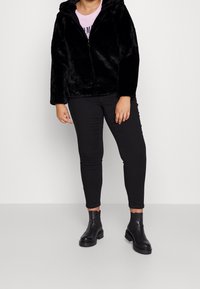 Zwarte nepbont ritsvest met capuchon, lange mouwen en een ontspannen pasvorm, gecombineerd met zwarte slimfit broek en zwarte Chelsea-laarzen.