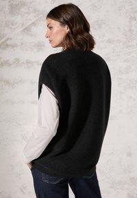 Sort ribbet strikket ærmeløs sweater med en afslappet pasform, båret over en hvid langærmet skjorte, matcher med mørk denim jeans.