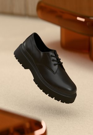 Black leather lace-up shoes with textured rubber sole, featuring a rounded toe and low-profile design, suspended above a beige surface.
