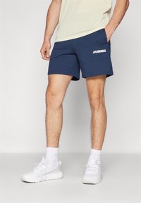 Marinblåa träningsshorts med elastisk midja. Har en vit "HUMMEL"-logotyp på vänster sida. Tyget har en jämn textur.